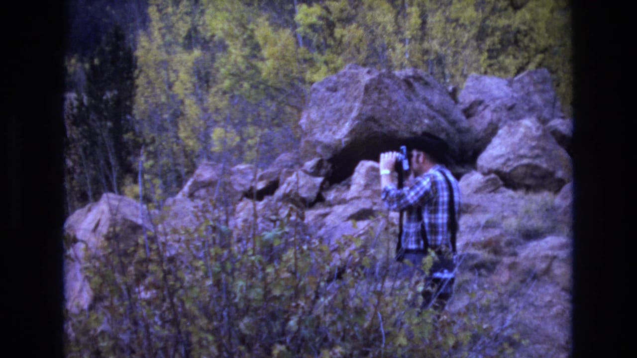 In 1973 Colorado Springs 8mm footage, an adult man in a plaid shirt and dark hat or hair profile looks through binoculars beside large rocks and scrubby vegetation.