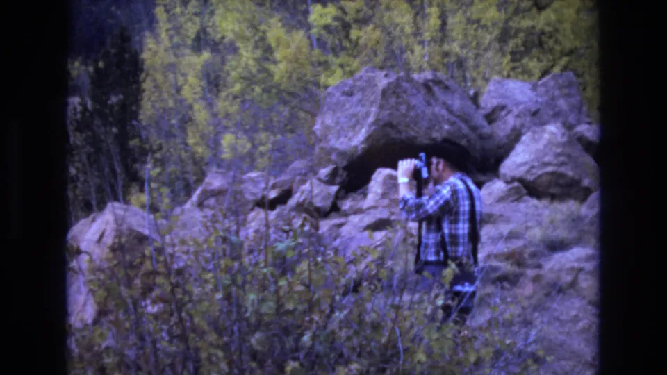 In 1973 Colorado Springs 8mm footage, an adult man in a plaid shirt and dark hat or hair profile looks through binoculars beside large rocks and scrubby vegetation.
