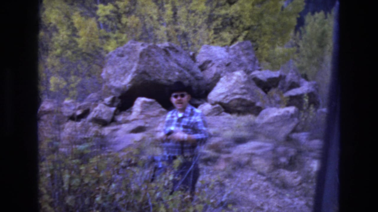 Man in sunglasses stands among boulders and brush holding gear.