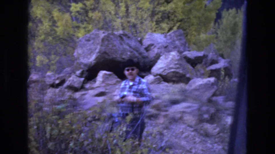 In a rocky Colorado Springs outdoor setting, the man faces the camera wearing sunglasses and a plaid shirt while standing amid brush with boulders behind him.