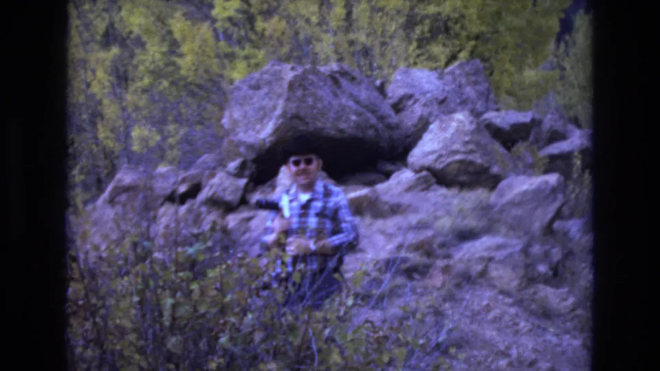 The 1973 home-movie frame shows the man partly obscured by foreground brush as he moves or pauses below large boulders in the Colorado Springs wilderness setting.
