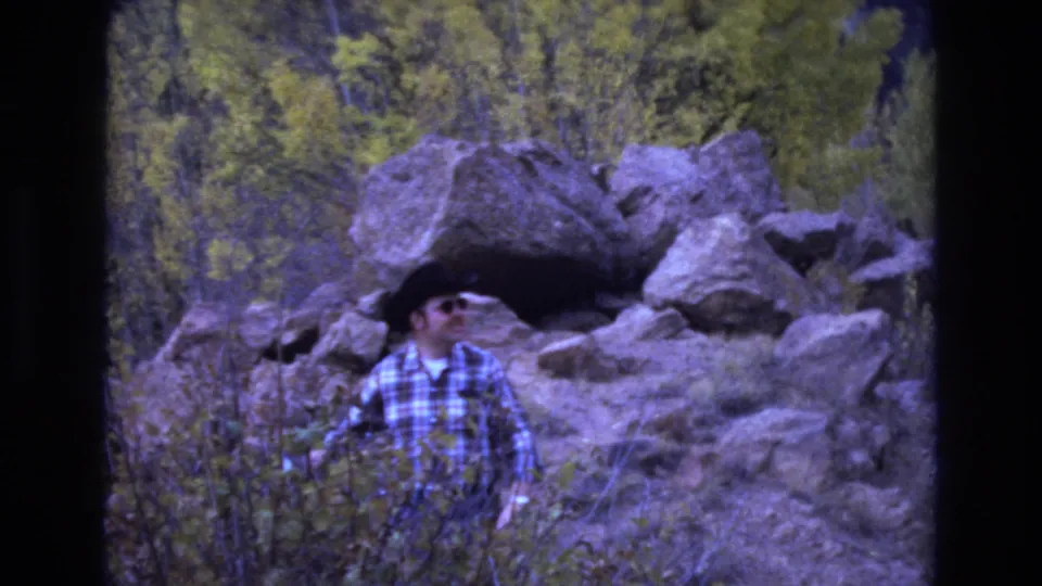 In the final frame, the man in sunglasses and plaid flannel looks off to one side while standing in scrubby vegetation with large boulders and trees behind him.
