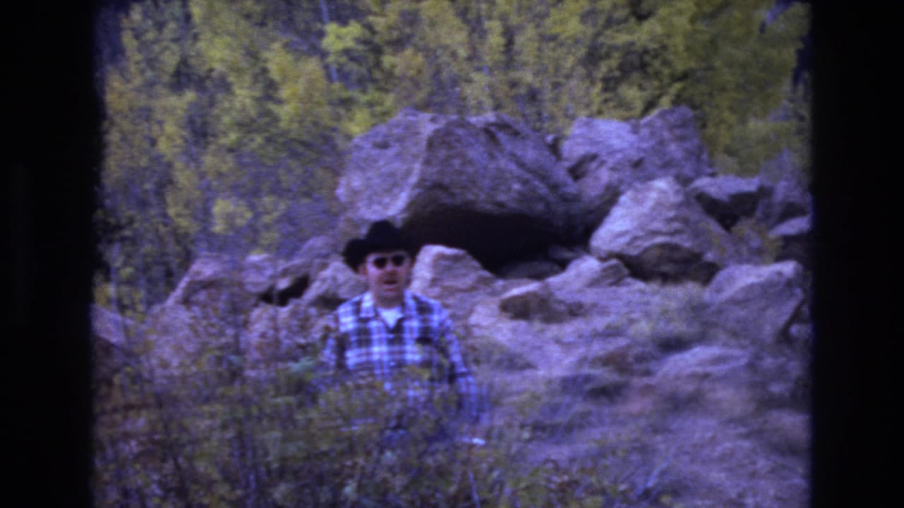 In 1973 Colorado Springs footage, an adult man in a black cowboy hat, sunglasses, and plaid shirt stands among dry brush with large boulders and trees behind him.
