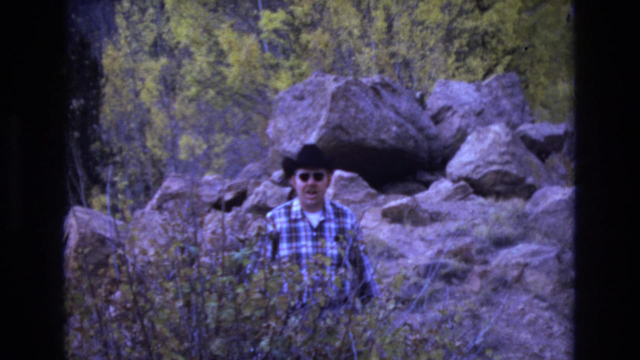 Man in cowboy hat facing the camera with boulders and brush behind him.