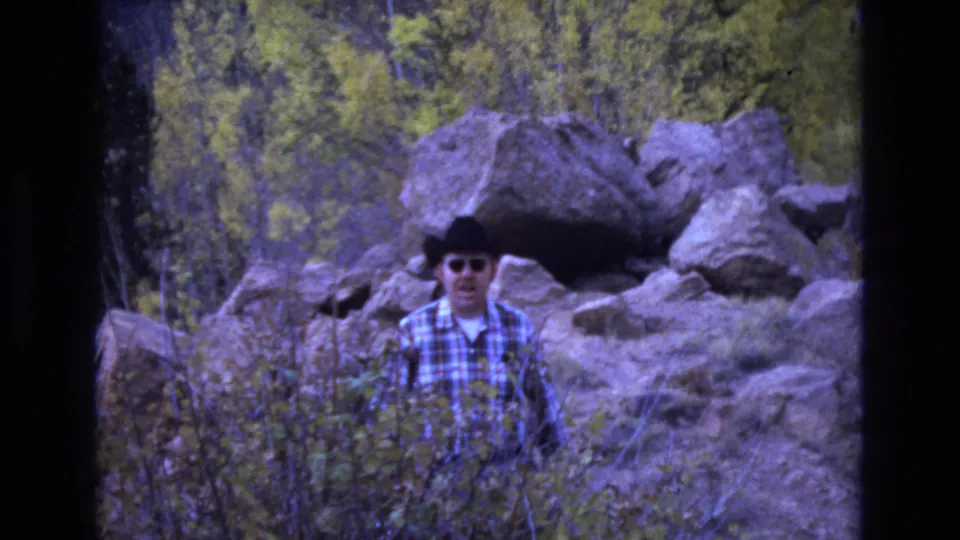 The man faces toward the home-movie camera in the Colorado Springs landscape, wearing sunglasses and a plaid shirt amid scrubby plants and large rocks.