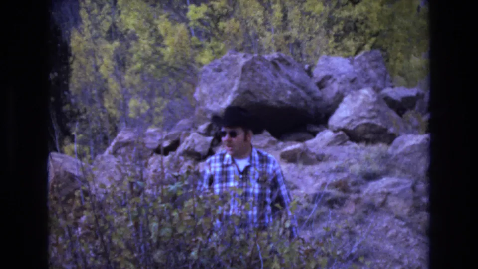 The man turns slightly to one side in the 1973 home-movie frame, partly screened by brush with boulders and yellow-green trees in the background.