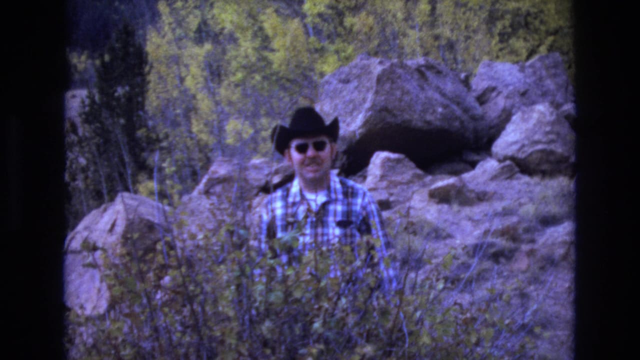 Adult man in a black cowboy hat, sunglasses, and blue plaid shirt stands among rocks and brush in Colorado Springs home-movie footage from 1973.
