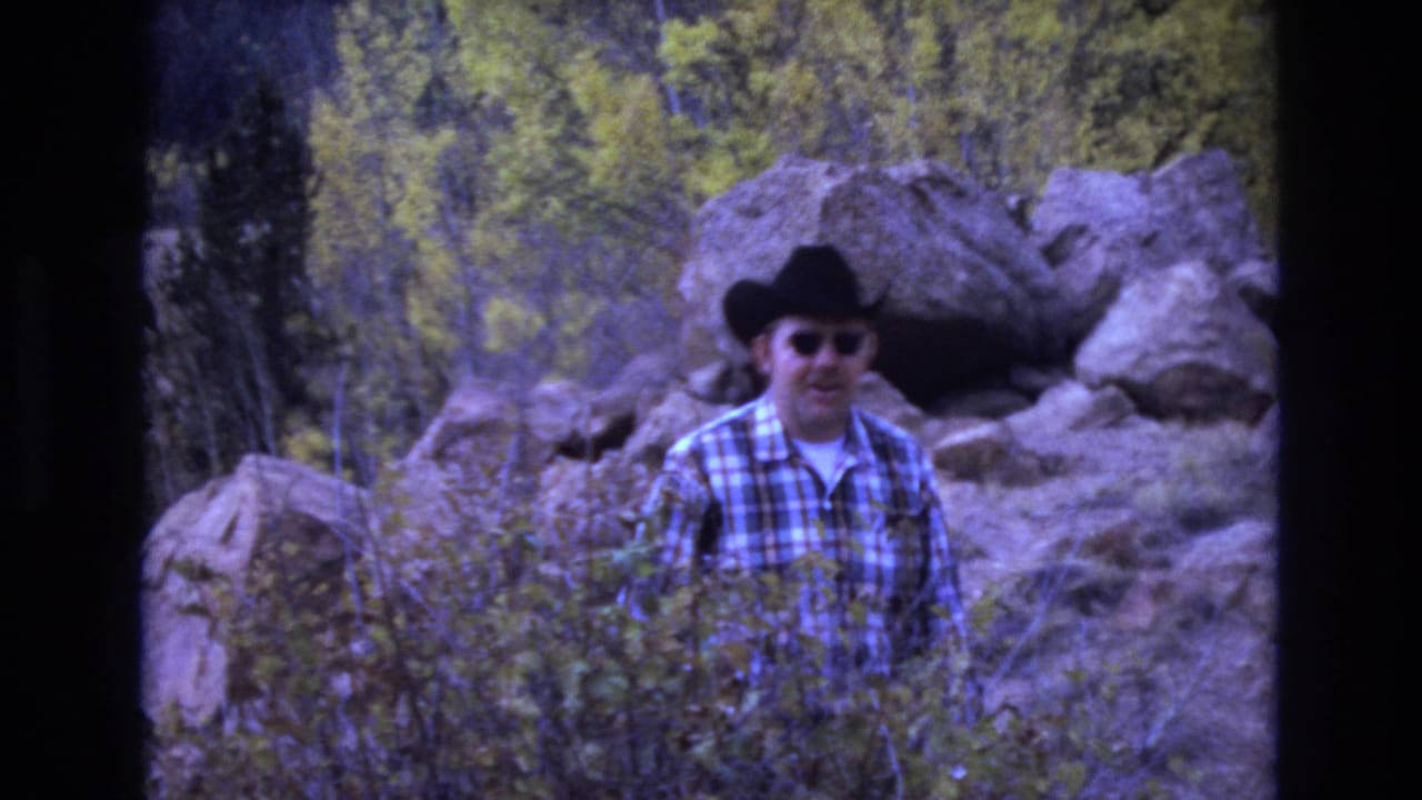 Man in a cowboy hat standing in front of boulders and autumn foliage.