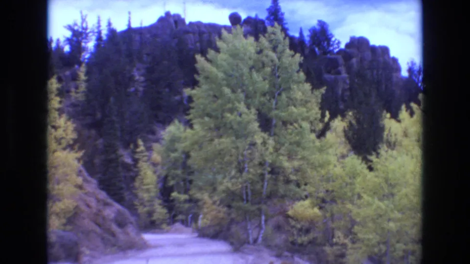 A winding mountain road passes through yellow-green aspens and dark evergreens with a rocky ridgeline beyond, filmed on 8mm in the Colorado Springs area in 1973.