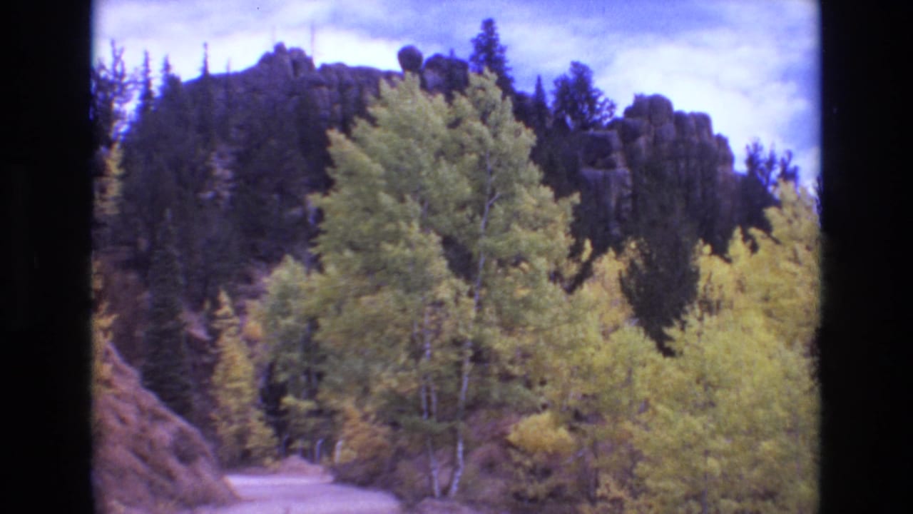 1973 Colorado Springs home-movie frame showing a dusty mountain road curving between yellow autumn trees, evergreen forest, rocky slopes, and distant hills.