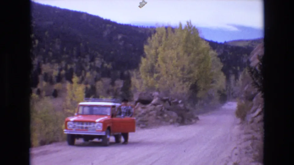 1973 Colorado Springs home-movie frame showing the red truck closer to camera on a dusty mountain road, with the driver's door open and an adult figure partly visible beside it.