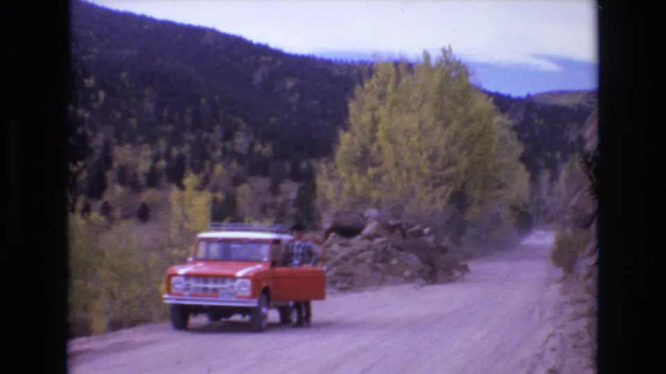 1973 Colorado Springs frame showing a red truck parked near the left side of a dusty mountain road, with an adult man by the open door and autumn trees behind.