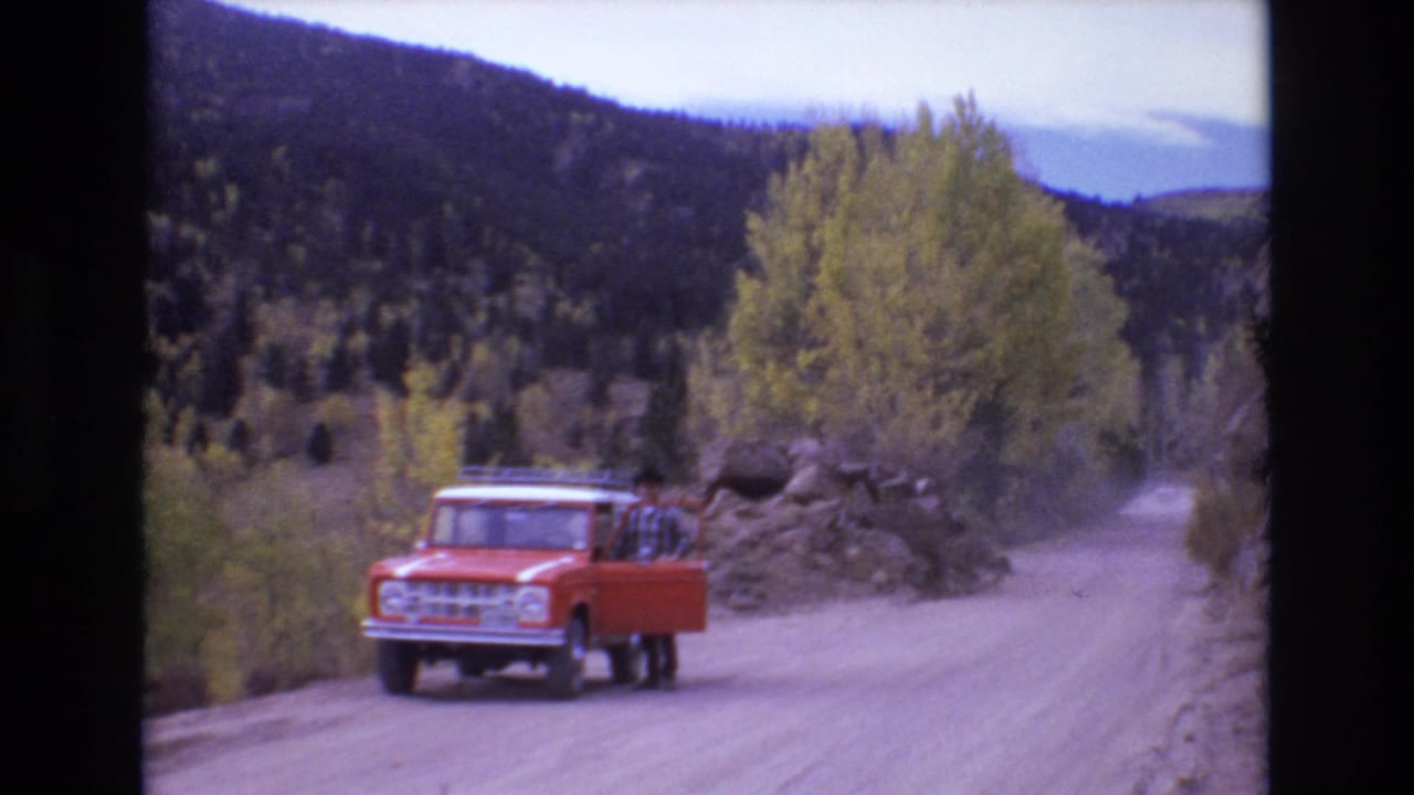 1973 Colorado Springs home-movie frame showing an orange utility truck on a dusty mountain road while a man in plaid stands by the open driver-side door.