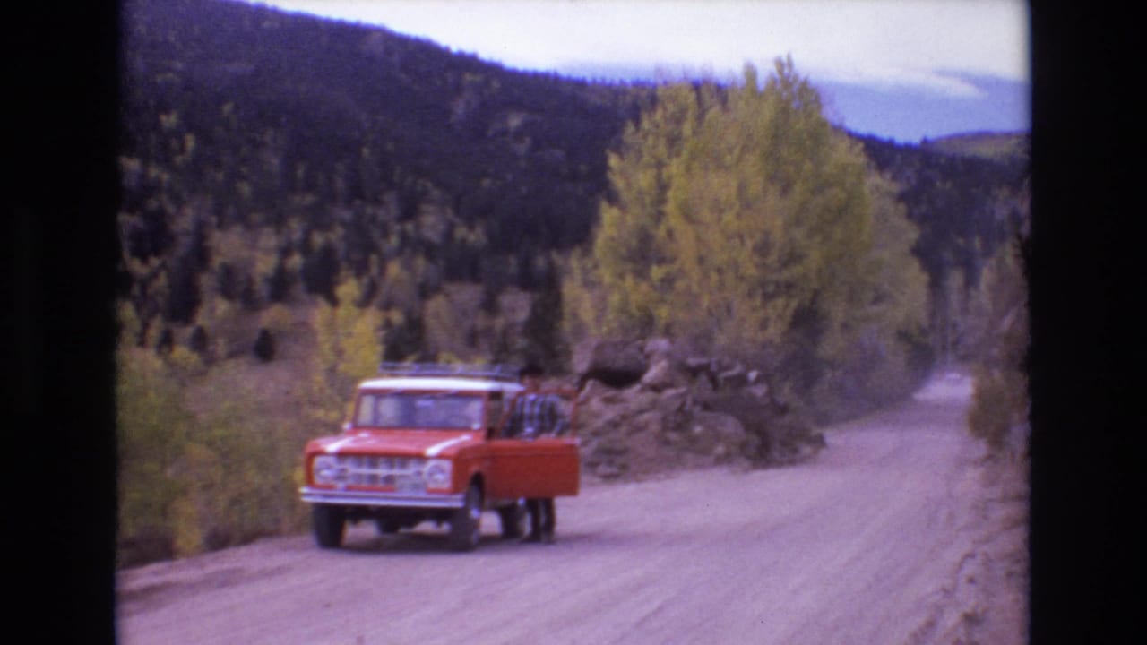 Man stands at the open door of an orange truck parked on a curving dirt road.