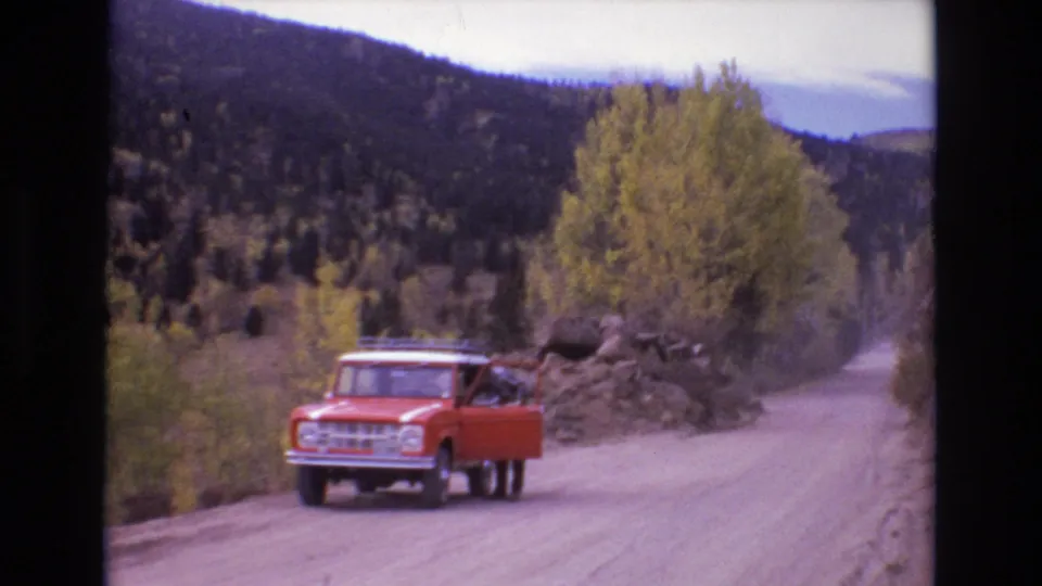 1973 Colorado Springs home-movie still showing an orange truck with an open driver-side door on a winding dirt road below wooded hills.