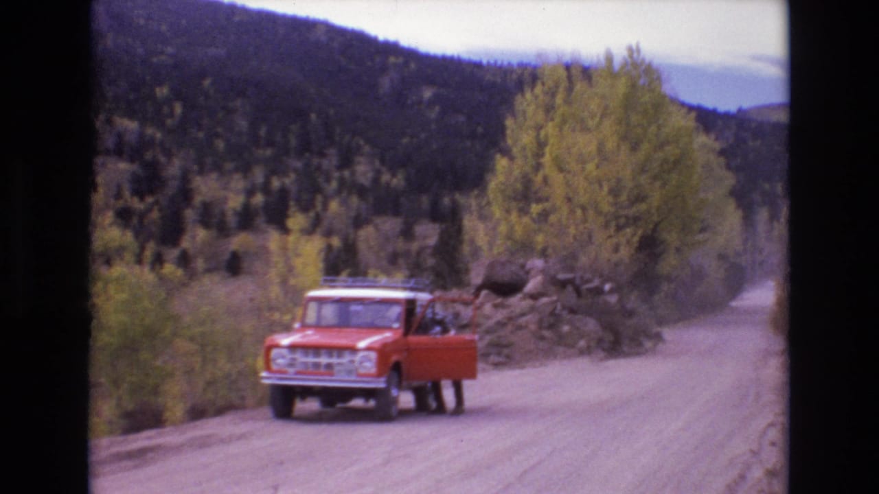 1973 Colorado Springs home-movie frame showing a red classic Ford Bronco on an unpaved mountain road, with a person near the open door and wooded hills behind.