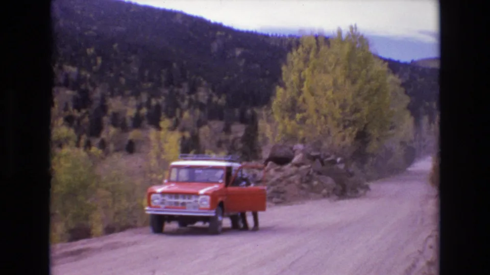 1973 Colorado Springs home-movie frame showing a red classic Ford Bronco on an unpaved mountain road, with a person near the open door and wooded hills behind.