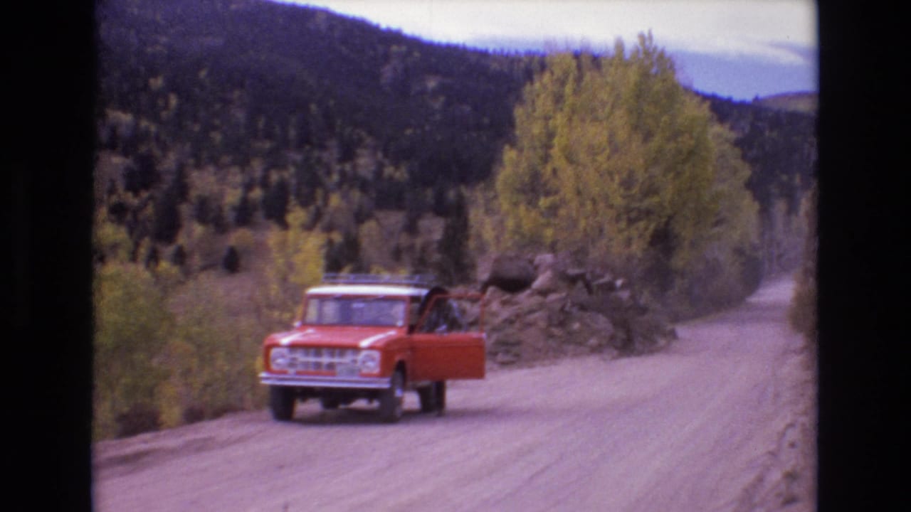 Red vehicle on a curving dirt road with its side door open near trees and rocks.