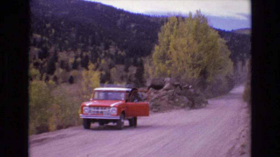 The red Ford Bronco appears on a dusty Colorado mountain road with an open door, roadside rocks, autumn foliage, and forested hills in the background.