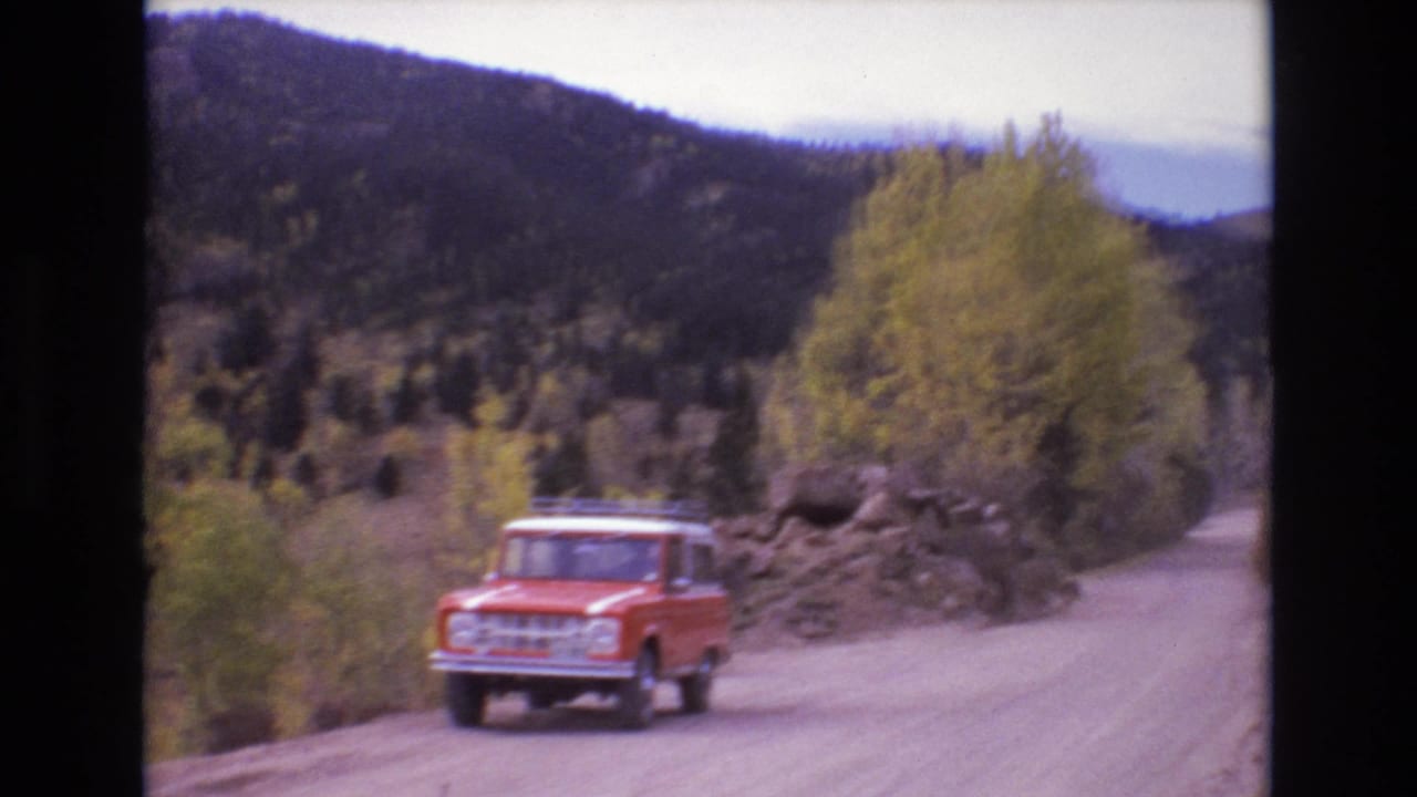 A red older SUV drives along an unpaved mountain road in Colorado Springs, with forested hills and autumn-colored trees in the background.