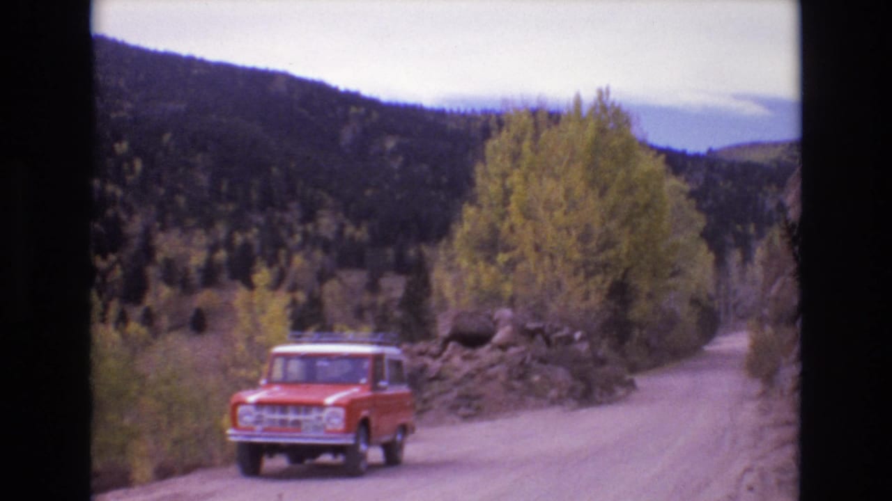 The red SUV approaches on a pale dirt road bordered by yellow trees, roadside rock piles, and dark wooded hills in the Colorado Springs area.