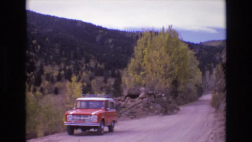 The red SUV approaches on a pale dirt road bordered by yellow trees, roadside rock piles, and dark wooded hills in the Colorado Springs area.