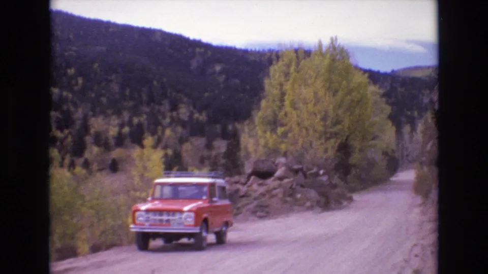 A red older SUV is framed nearer to the camera on a dusty Colorado mountain road, with trees, rocks, and layered hills behind it.