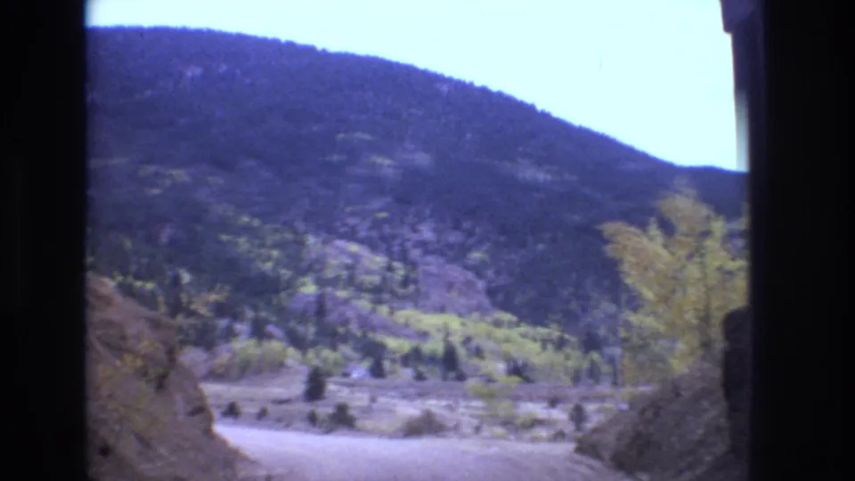 Colorado Springs travel footage showing a bright roadside view of a forest-covered mountain, pale dirt road, rocky bank, and scattered yellow-green trees.