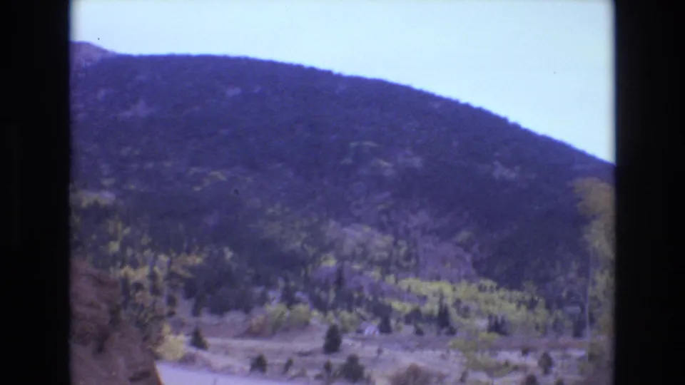 Vintage 8mm frame of a Colorado mountain landscape with a winding dirt road, wooded slope, open ground, and autumn-toned trees under bright sky.