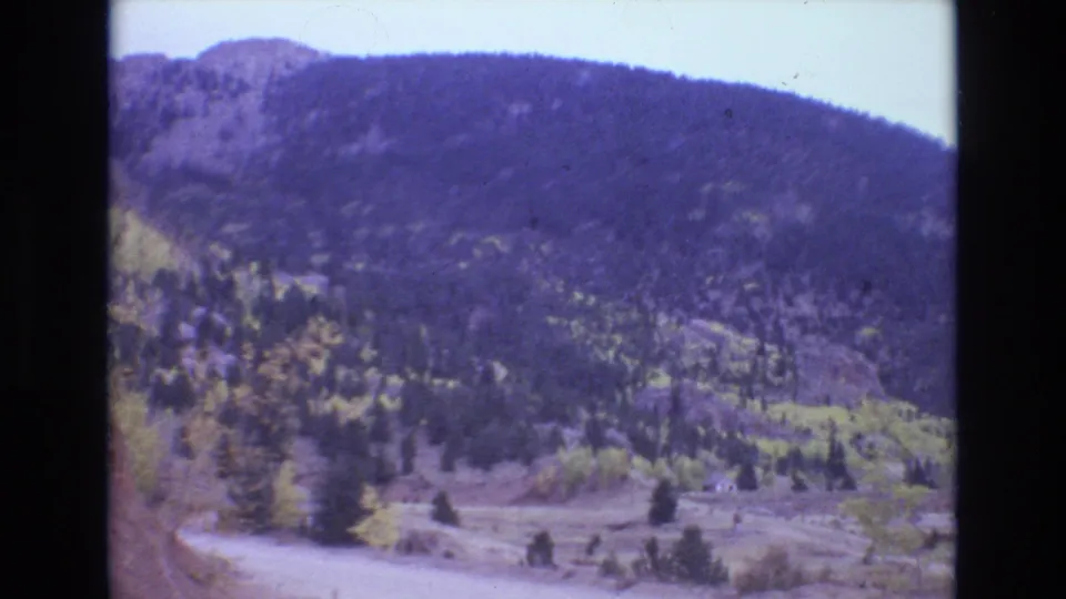 1973 Colorado Springs home-movie view of a forested mountain valley with a pale road, rocky roadside cut, scattered trees, and blue-shifted Super 8 color.