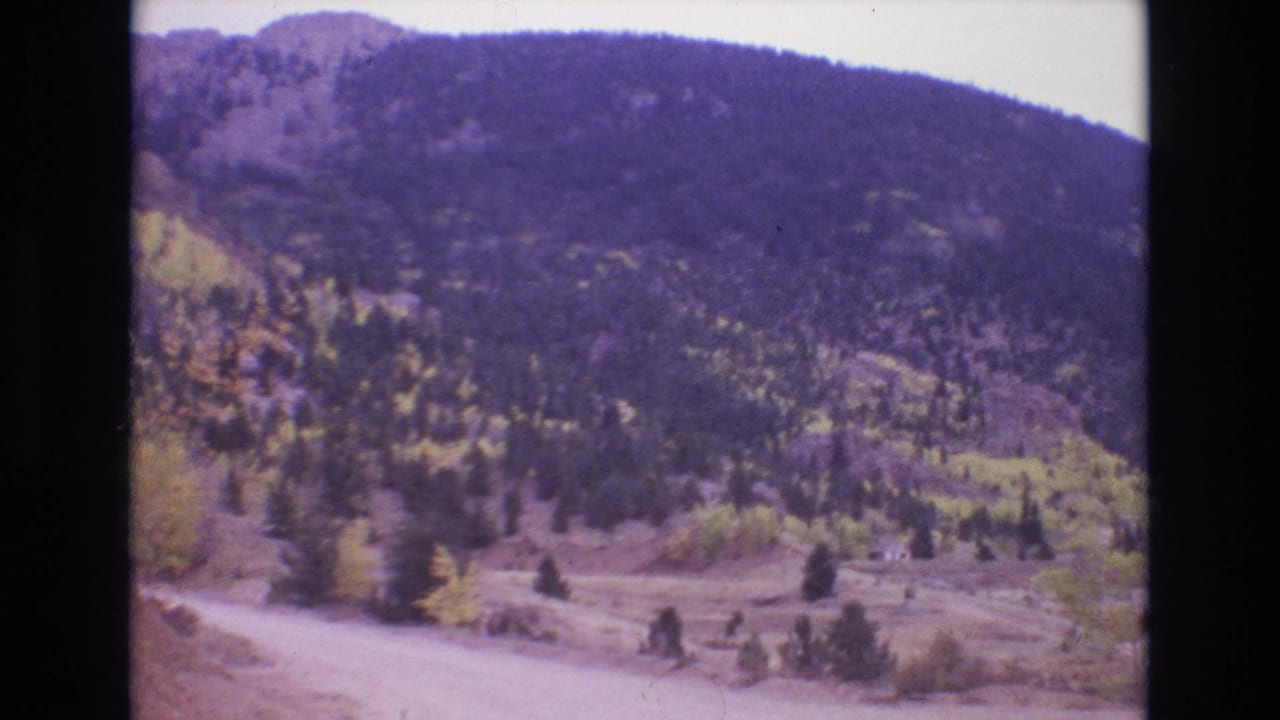 1973 Colorado Springs-area 8mm frame showing a pale dirt road at the base of a forested mountain slope with evergreen trees and yellow autumn foliage.