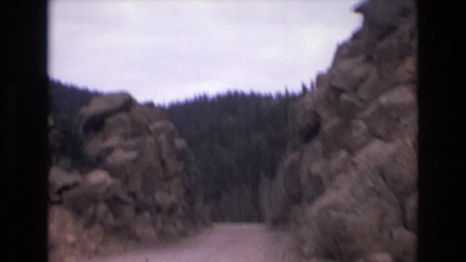 1973 Colorado Springs-area 8mm frame showing a dirt road cutting between large rocky outcrops, with evergreen forest and pale overcast sky beyond.