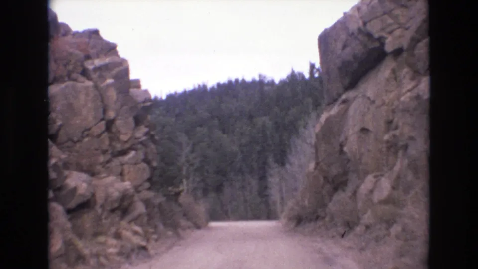 1973 Colorado Springs home-movie frame of an unpaved road bordered by tall canyon rocks with pine forest beyond.