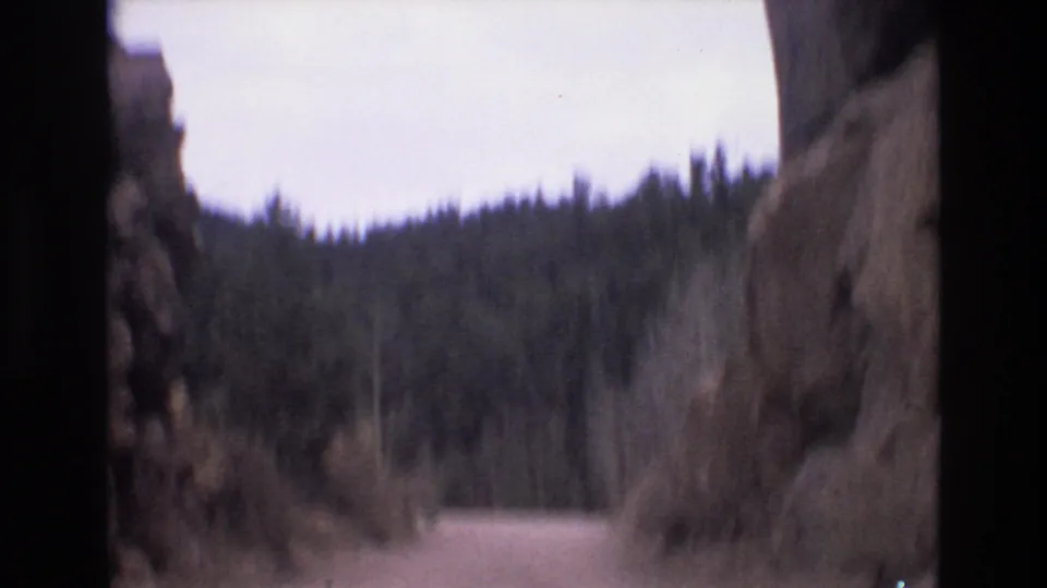 Soft 8mm frame from the Colorado Springs road sequence, with roadside rock close to camera and a pine-covered slope ahead.
