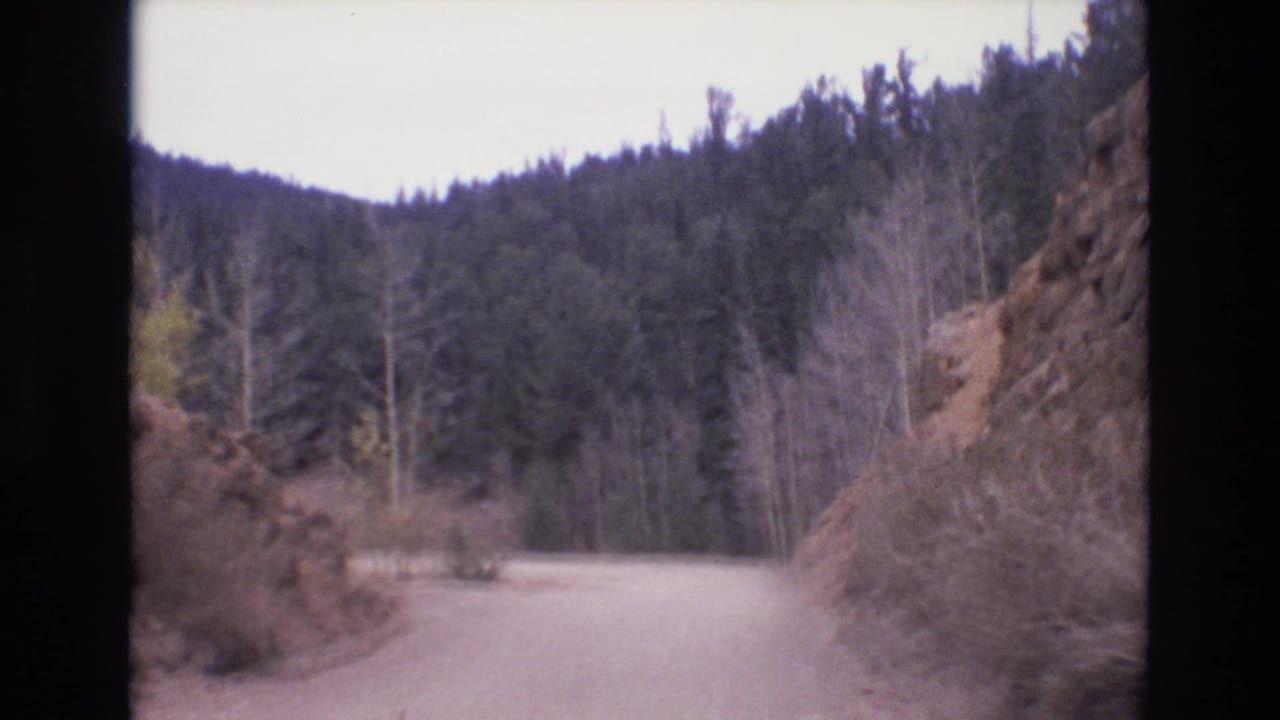 1973 Colorado Springs 8mm frame showing a pale dirt road bending through forested mountain terrain with rocky cuts and distant wooded slopes.