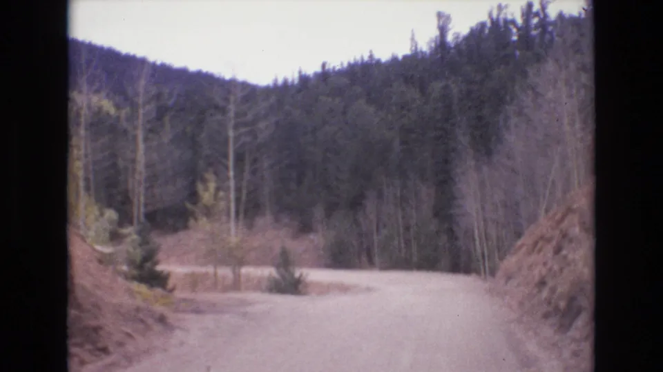 1973 Colorado Springs home-movie frame with an unpaved road curving through evergreen and bare trees below dark mountain slopes.