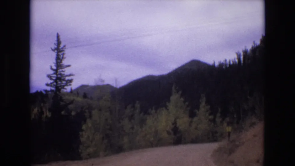 1973 Colorado Springs 8mm frame showing the road foreground opening toward layered wooded mountain ridges under a cloudy sky.