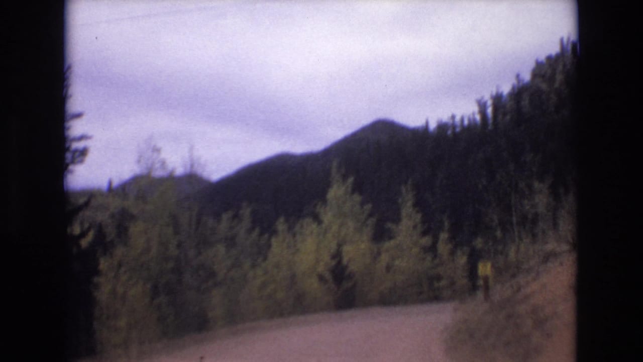 1973 Colorado Springs home-movie frame showing a pale winding road at the edge of dense forest with dark mountain slopes under an overcast sky.