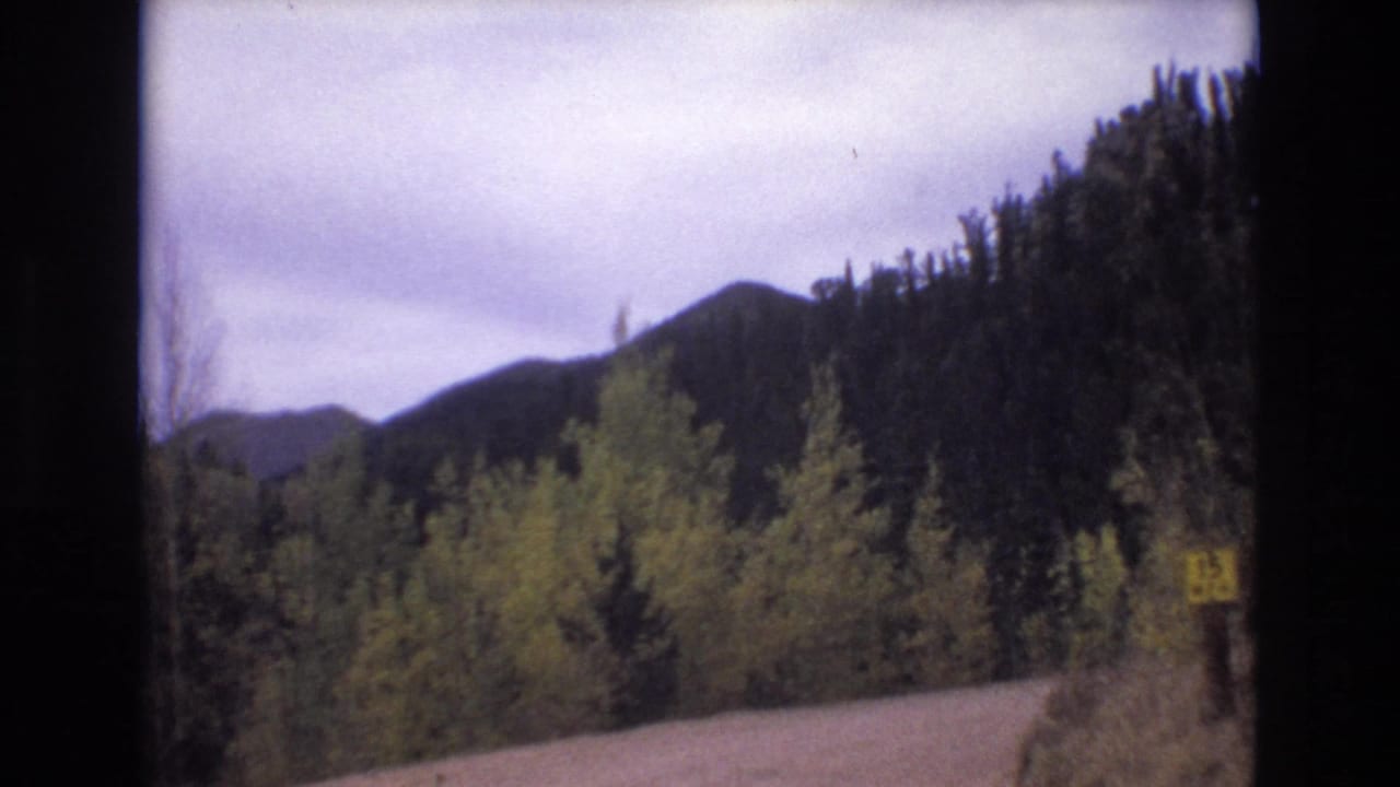 1973 Colorado Springs frame with a road bending through wooded terrain, a yellow roadside sign, and layered mountain ridges in the distance.
