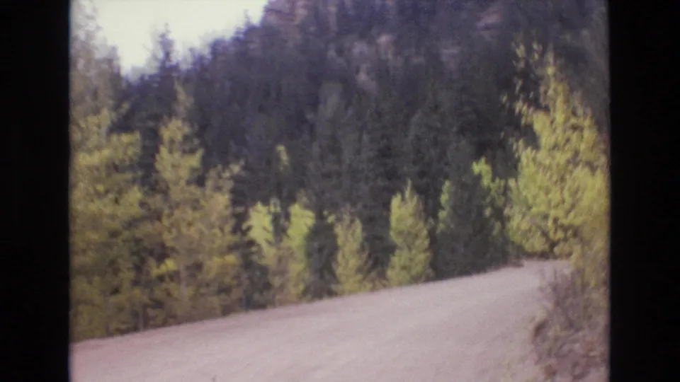 1973 Colorado Springs home-movie frame looking along a pale road bordered by dense trees and a shadowed forested slope.