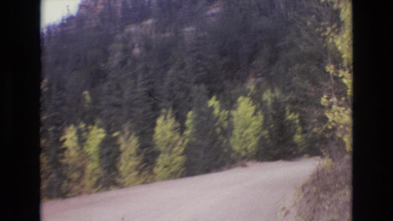 1973 8mm home-movie frame from the Colorado Springs area showing a pale unpaved road bending through evergreen and yellow-green hillside foliage.