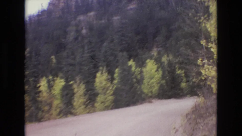 1973 8mm home-movie frame from the Colorado Springs area showing a pale unpaved road bending through evergreen and yellow-green hillside foliage.