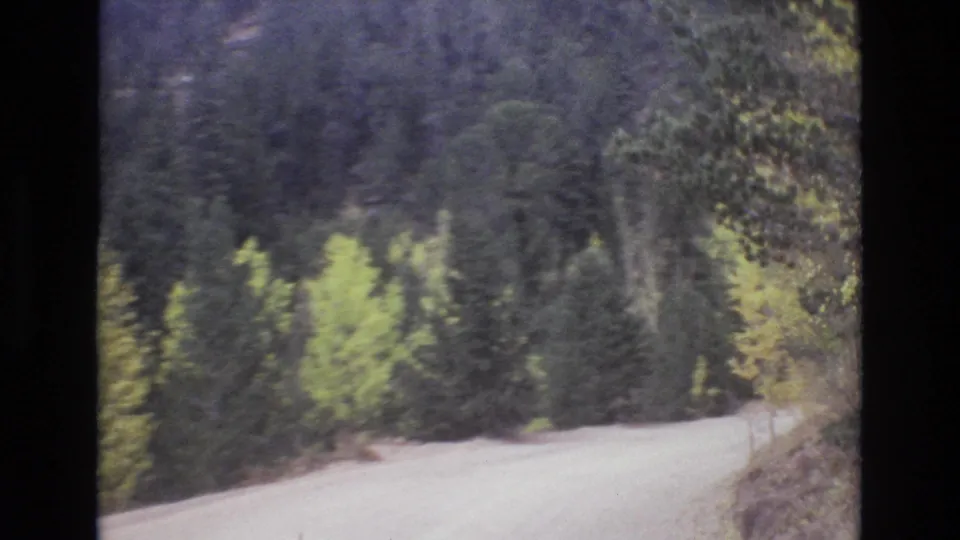 Colorado Springs area travel footage from 1973 shows an unpaved mountain road edging past dark evergreens and patches of yellow-green trees.