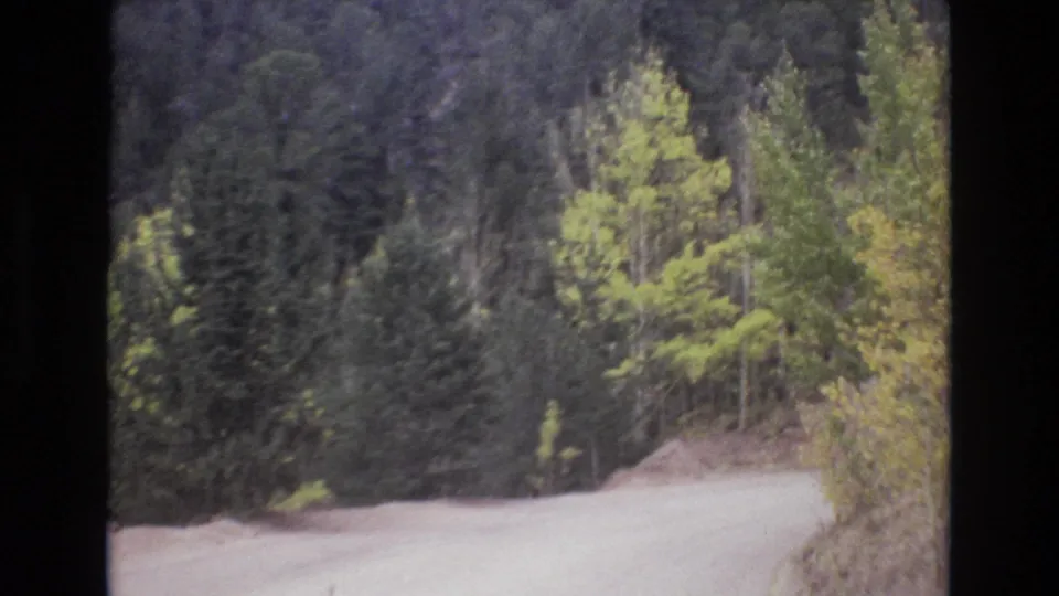 A 1973 8mm frame shows the road turning along a wooded hillside near Colorado Springs, with thick evergreens, brush, and muted home-movie color.