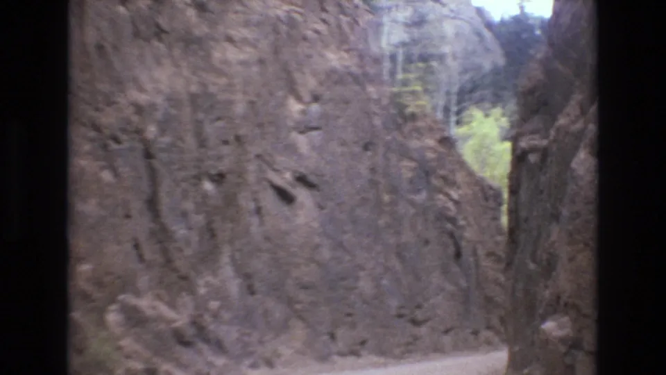 The camera passes a steep rock wall on a dirt road, with a tree trunk and wooded background visible in the 1973 Colorado Springs travel sequence.