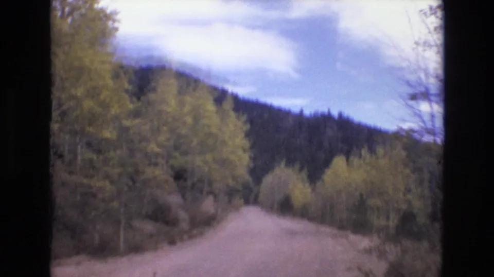 1973 8mm home-movie frame from the Colorado Springs area showing the road ahead, yellowing roadside trees, forested hills, and a bright clouded sky.