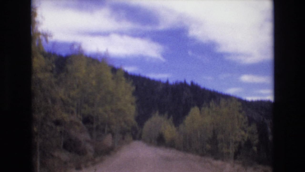 1973 8mm home-movie frame from the Colorado Springs area showing a narrow dirt road bordered by trees and dark mountain slopes under scattered clouds.