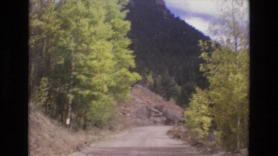 1973 8mm home-movie frame showing a curving unpaved road, bright green trees, and a rocky mountainside in the Colorado Springs area.