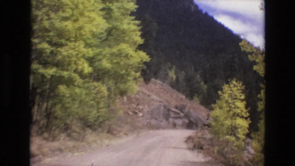 1973 8mm home-movie frame from the Colorado Springs area showing a dirt road curving through trees with a rocky cut bank and mountain ridge beyond.