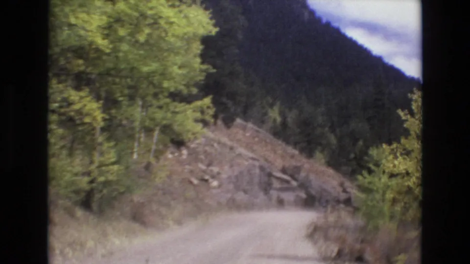 1973 8mm home-movie frame showing a narrow dirt road bending through a wooded mountain landscape near Colorado Springs, with brush and a dark hillside beyond.